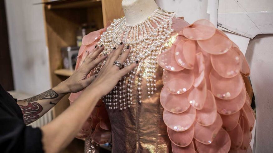 Créatrice en train de positionner un collier de perles sur un costume de carnaval aux épaules ornées de pétales rose saumon brillants, dans un atelier de confection.
