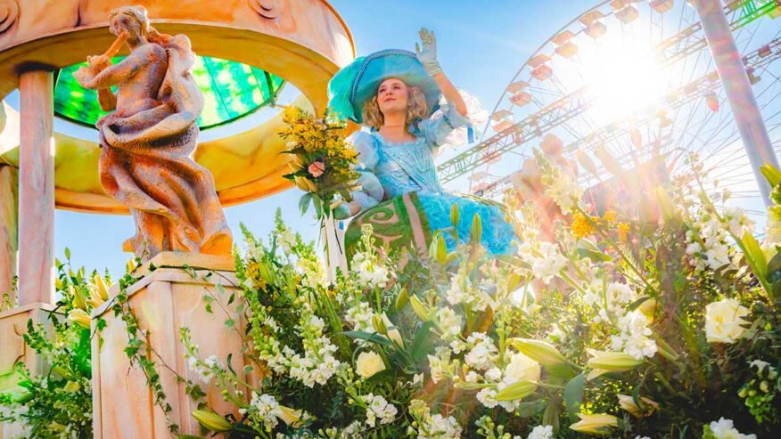 Char décoré de fleurs lors de la Bataille de Fleurs du Carnaval de Nice. Une femme costumée en tenue d’époque bleue, coiffée d’un large chapeau, salue la foule depuis le char, avec en arrière-plan une grande roue et un soleil éclatant.