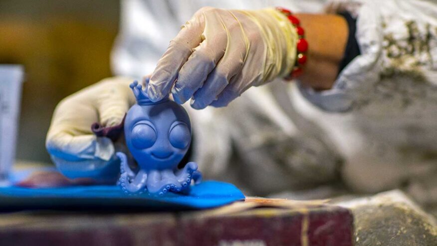 Gros plan sur les mains gantées d’un carnavaliers travaillant une figurine bleue en forme de pieuvre couronnée, dans un atelier du Carnaval de Nice.