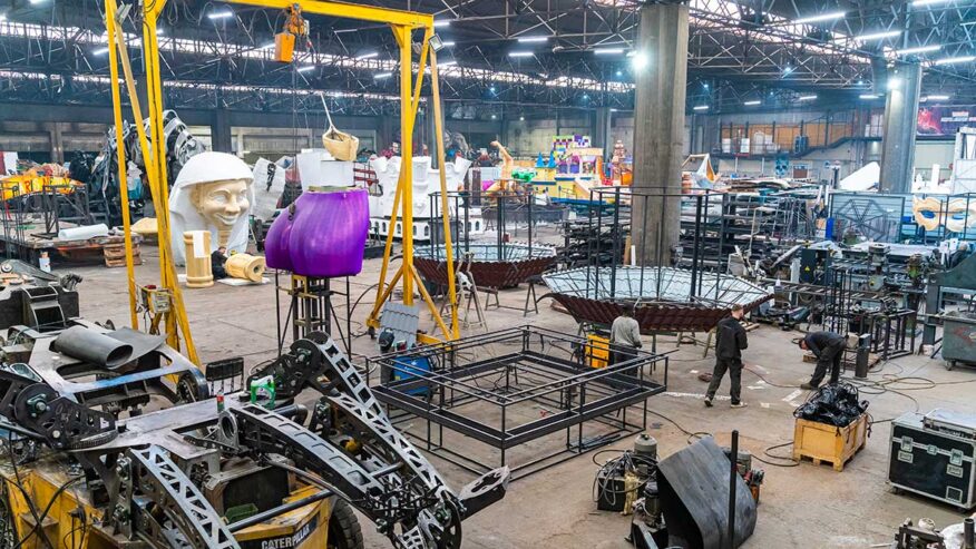 Atelier de construction de chars du Carnaval de Nice dans la grande Halle Spada. Plusieurs structures métalliques en cours de fabrication, éléments décoratifs colorés, et carnavaliers au travail. En arrière-plan, une grande tête de personnage en résine blanche.